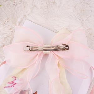 Puede incluir: Un gran lazo rosa y crema con una pinza de cocodrilo plateada. El lazo est&aacute; hecho de tela transparente y tiene un aspecto delicado y femenino.