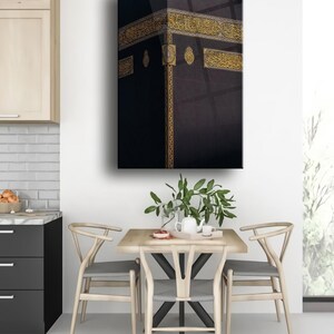 May include: A black and gold framed canvas print of the Kaaba, a cube-shaped building in Mecca, Saudi Arabia. The Kaaba is covered in a black cloth with gold embroidery and Arabic calligraphy.