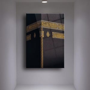 May include: A black and gold framed canvas print of the Kaaba, a cube-shaped building in Mecca, Saudi Arabia, that is Islam's holiest place. The Kaaba is covered in black cloth and has gold trim. The print is framed in a black frame.