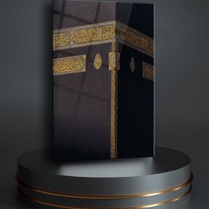May include: A black and gold framed canvas print of the Kaaba, a cube-shaped building in Mecca, Saudi Arabia. The Kaaba is covered in black cloth and has gold trim. The print is on a gray pedestal with gold accents.