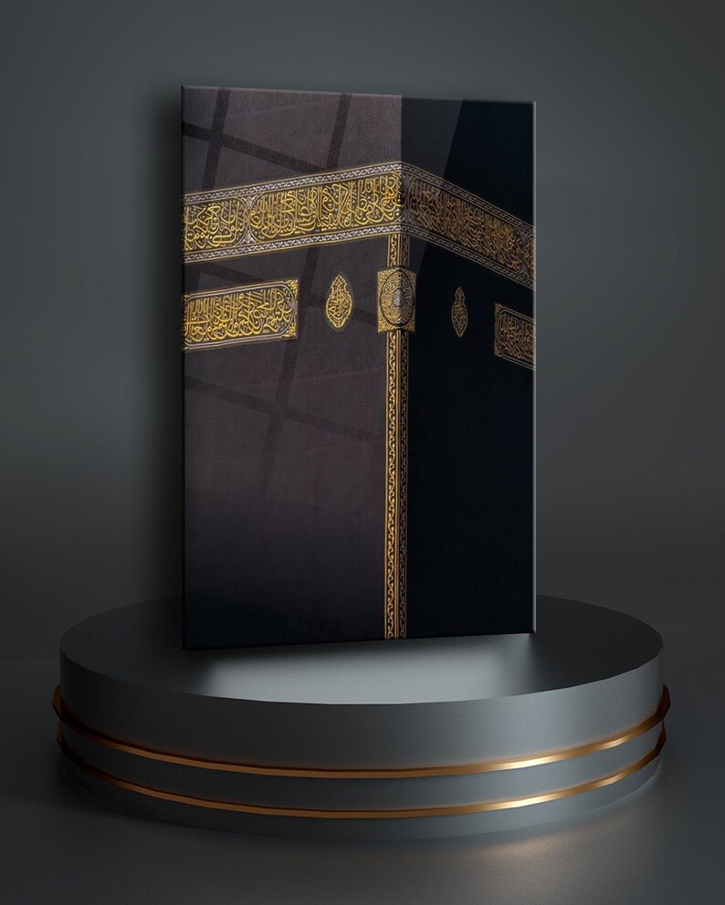 May include: A black and gold framed canvas print of the Kaaba, a cube-shaped building in Mecca, Saudi Arabia. The Kaaba is covered in black cloth and has gold trim. The print is on a gray pedestal with gold accents.