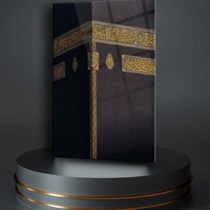 May include: A black and gold framed canvas print of the Kaaba, a cube-shaped building in Mecca, Saudi Arabia. The Kaaba is covered in black cloth and decorated with gold inscriptions in Arabic.