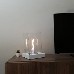 May include: A white tabletop fireplace with a glass enclosure and a flickering flame. The fireplace is on a dark brown wooden table with a small green plant in a white pot.