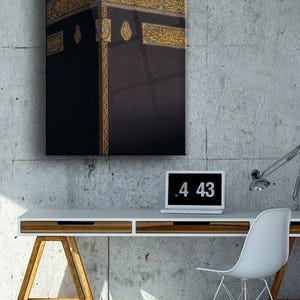 May include: A black and gold framed print of the Kaaba, a cube-shaped building in Mecca, Saudi Arabia, which is Islam's holiest place. The print is hanging on a concrete wall above a white desk with a laptop and a lamp.