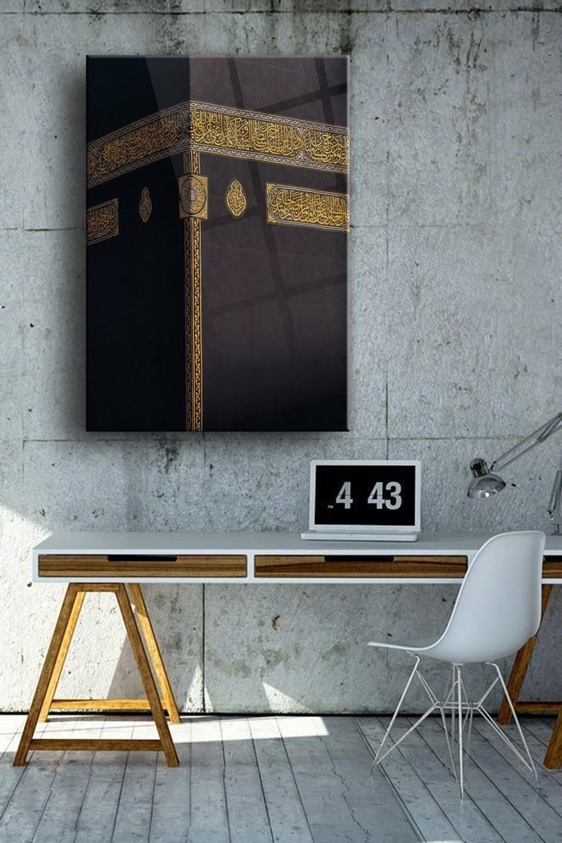 May include: A black and gold framed print of the Kaaba, a cube-shaped building in Mecca, Saudi Arabia, which is Islam's holiest place. The print is hanging on a concrete wall above a white desk with a laptop and a lamp.