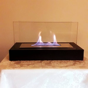 Op de afbeelding: Een tafelhaard met een zwarte basis en een transparante glazen omhulling. De haard heeft een zilveren metalen brander en blauwe vlammen.