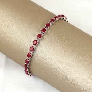May include: A silver bracelet featuring a series of oval-cut, vibrant red gemstones. The gemstones are set closely together, creating a continuous line of color. The bracelet is displayed on a neutral, textured background.