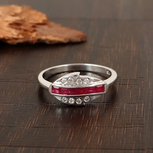 May include: A silver ring featuring a central oval design. The oval is filled with a row of small, square, red gemstones, and is bordered by small, round, clear gemstones. The ring is set against a dark wood surface.