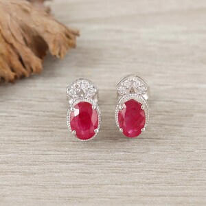 Puede incluir: Un par de pendientes de plata con piedras preciosas rojas oscuras de forma ovalada. Cada pendiente tiene un halo de pequeñas piedras claras alrededor del rubí y una parte superior decorativa con más piedras claras. Los pendientes se muestran sobre una superficie de vetas de madera clara.