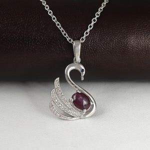Peut inclure: Un collier pendentif en argent en forme de cygne, orné d'un rubis et de petites pierres claires sur l'aile. Le cygne a un œil en pierre bleue et est suspendu à une fine chaîne en argent. Le collier est présenté sur une surface blanche.