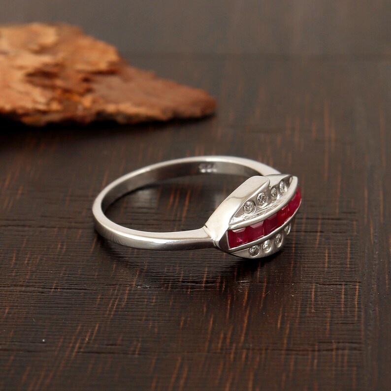 May include: A silver ring featuring a row of red gemstones and clear accent stones. The ring has a polished band, and the gemstones are set in a decorative setting. The ring is presented on a dark wooden surface.