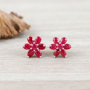 May include: A pair of flower-shaped stud earrings. Each earring features a cluster of oval-cut, deep red gemstones arranged around a central stone. The settings are silver-colored, and the earrings are displayed on a light wooden surface.