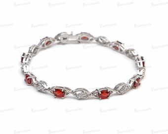 May include: A silver bracelet with alternating oval-cut red gemstones and small, round, clear gemstones. The bracelet is linked with a delicate chain and has a secure clasp. The brand name "Reevaans" is visible in the background.