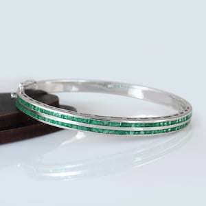 May include: A silver-toned hinged bangle bracelet featuring two parallel rows of square-cut green gemstones set along the outer edge. The bracelet has a polished metallic finish and a secure clasp mechanism visible on the left side of the frame.