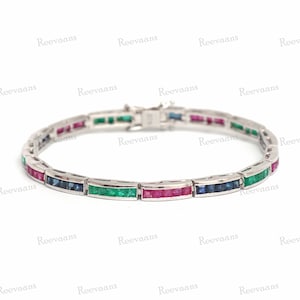 Pode incluir: Uma pulseira de prata com uma fila de pedras preciosas de esmeralda, rubi e safira. A pulseira é feita de metal branco e tem um fecho.