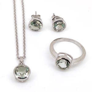 Könnte beinhalten: Ein Schmuckset mit einer Halskette, Ohrringen und einem Ring aus Silber. Jedes Stück hat einen runden, hellgrünen Edelstein, der in einer silbernen Lünette gefasst ist. Die Halskette hat eine feine Kette.