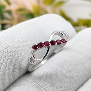 Könnte beinhalten: Ein silberfarbener Ring mit einem Unendlichkeits-Symbol-Design. Der Ring hat eine Reihe von fünf kleinen, runden, tiefroten Edelsteinen. Der Ring ist vor einem weißen Hintergrund platziert, was den Kontrast zwischen dem Metall und den Edelsteinen hervorhebt.