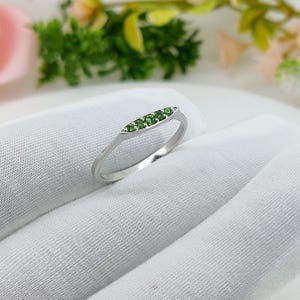 May include: A silver ring with a row of five small, round, bright green gemstones set in a horizontal bar. The ring band is thin and polished. The ring is displayed on a white, textured surface.