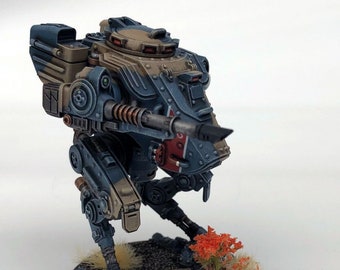May include: A blue and tan painted miniature war machine with a gun barrel and a red stripe on the side. The machine is standing on a round base with brown and green grass.