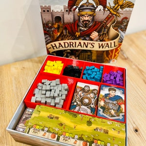 Puede incluir: Caja del juego de mesa "Hadrian's Wall" con una ilustración de un soldado romano. La caja abierta revela compartimentos rojos con piezas de juego amarillas, negras, azules y moradas, además de cartas y un tablero.