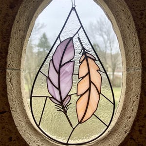 Pode incluir: Peça de arte em vitral com duas penas estilizadas, uma lavanda e outra pêssego, em forma de gota. A peça é emoldurada em preto e pendurada em uma janela oval com moldura de pedra. O fundo mostra uma cena externa desfocada.