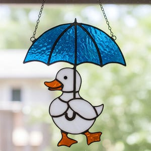 May include: A stained glass hanging featuring a white duck holding a blue umbrella. The duck has orange feet and a yellow beak. The umbrella has a textured glass design.