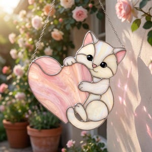 May include: A stained glass cat holding a pink heart. The cat is white with pink accents on its ears and nose. The heart has a marbled pink design. A silver chain is attached for hanging.