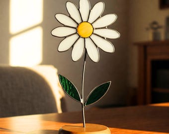 May include: A stained glass daisy flower with a yellow center and green leaves on a wooden base.