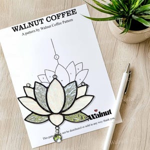 May include: A white paper with a stained glass lotus flower pattern, labeled "WALNUT COFFEE" and "A pattern by Walnut Coffee Pattern." A white pen rests beside the pattern. The lotus design features white and green glass with a clear crystal pendant.