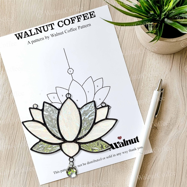 May include: A white paper with a stained glass lotus flower pattern, labeled "WALNUT COFFEE" and "A pattern by Walnut Coffee Pattern." A white pen rests beside the pattern. The lotus design features white and green glass with a clear crystal pendant.
