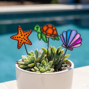Könnte beinhalten: Ein weißer Keramiktopf mit Sukkulenten, verziert mit dekorativen Gartensteckern. Die Stecker sind in Form eines Seesterns, einer Schildkröte und einer Muschel gehalten, in Orange, Grün und Lila. Der Hintergrund ist ein verschwommener blauer Pool.
