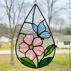 May include: Stained glass window art in a teardrop shape. The design features a pink flower with a yellow center, a light blue butterfly, and green leaves. The glass has a textured appearance and is framed with black lines.