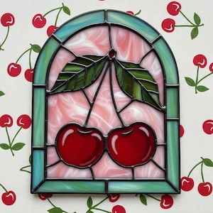 May include: Stained glass panel featuring a pair of bright red cherries with green leaves. The panel has a pink and white swirled background, framed by a teal and black border. The design is in an arched shape.