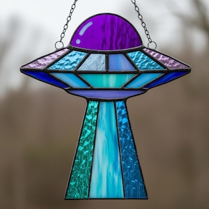 May include: A stained glass UFO suncatcher in shades of purple, blue, and teal. The alien spacecraft design is suspended by a chain. The glass has a textured appearance, with a mix of opaque and translucent sections.