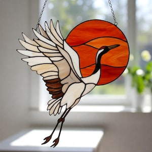 May include: A stained glass window hanging with a white crane flying in front of an orange sun. The crane has brown accents on its wings and a long, thin black beak.