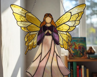 May include: A stained glass angel with yellow wings and a pink and purple dress. The angel is hanging from a chain.