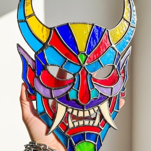 May include: A colorful stained glass mask in the shape of a demon's face. The mask features a variety of colors, including blue, yellow, red, and purple. It has horns, fangs, and a menacing expression. The mask is held by a hand.