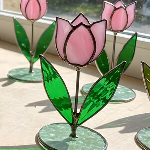 May include: Three pink stained glass tulips with green leaves and bases. The tulips are standing on a light wood surface.