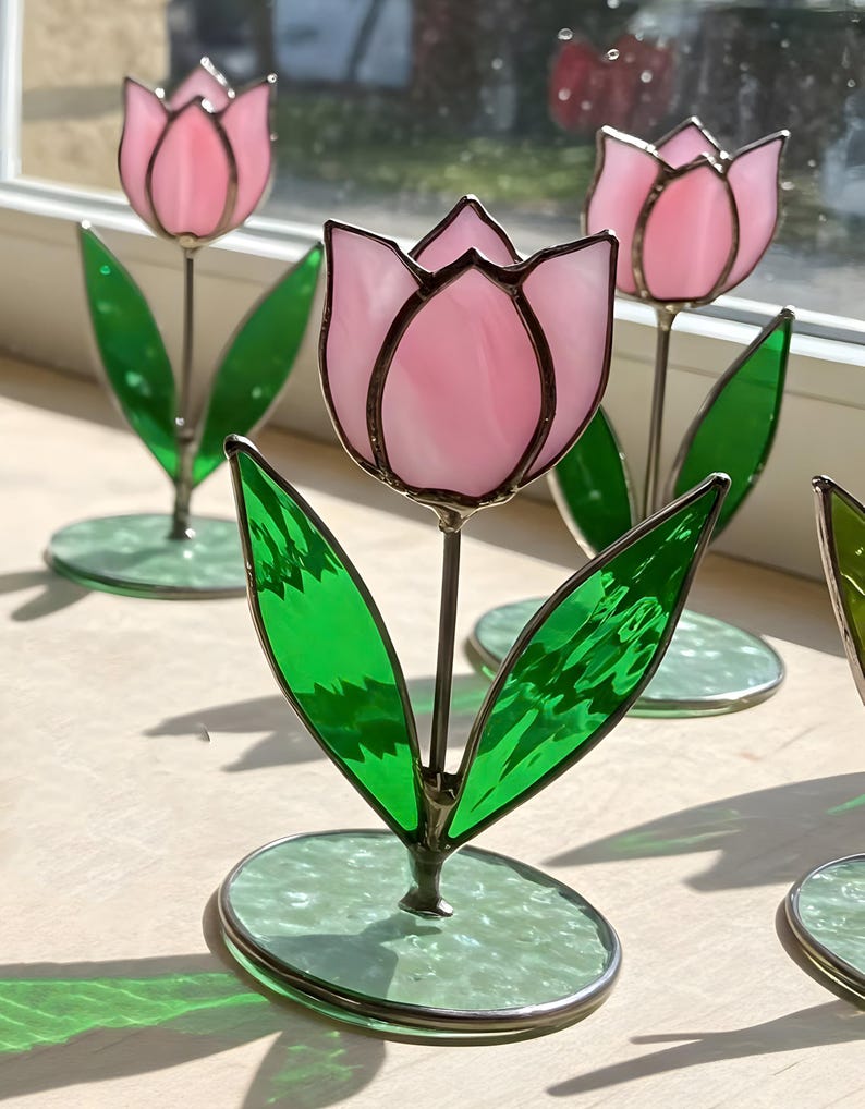 May include: Three pink stained glass tulips with green leaves and bases. The tulips are standing on a light wood surface.