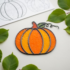 May include: A stained glass pumpkin in shades of orange and yellow, framed with black. The pumpkin features a stem and a curling vine. A pumpkin outline drawing is in the background, with green leaves.