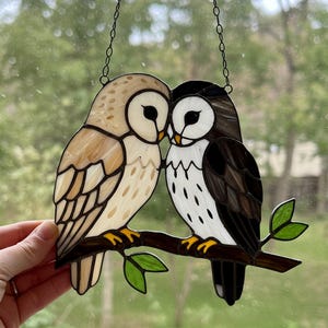 May include: A stained glass window hanging featuring two owls, one brown and white and the other black and white, perched on a branch with green leaves.