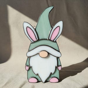 May include: A stained glass gnome with a green hat and pink bunny ears. The gnome has a white beard and a peach-colored nose.
