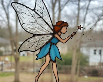 May include: A stained glass fairy with brown hair and a blue dress, holding a wand with a star on the end. The fairy is flying with her wings spread out.