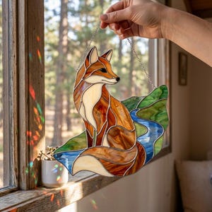 Puede incluir: Atrapasoles de vidrio con un zorro en tonos marrones, naranjas y crema, sentado sobre un paisaje verde y azul. La obra de arte se cuelga de una cadena y se muestra cerca de una ventana.