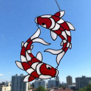 Puede incluir: Atrapasoles de peces koi de vidrieras. Dos peces koi rojos y blancos, conectados por una cadena, están suspendidos contra un cielo azul. Los peces tienen aletas y escamas detalladas, creando una pieza decorativa y artística.