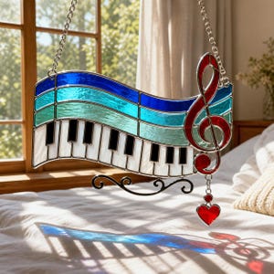 May include: Stained glass piano and treble clef suncatcher. The piano keys are white and black, with a blue and teal background. A red treble clef and heart charm hang below. The suncatcher is suspended by a silver chain.