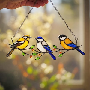 Op de afbeelding: Een hangend glas-in-loodraam met drie vogels die op een tak zitten met groene bladeren en oranje bessen. De vogels zijn een gele vogel, een blauwe vogel en een zwart-witte vogel.