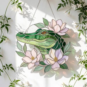 Può includere: Un acchiappasole in vetro colorato raffigurante una testa di alligatore verde con un occhio giallo, circondata da fiori di magnolia rosa chiaro e lavanda e foglie verdi. L'oggetto è appeso a una sottile catena metallica su una parete bianca.