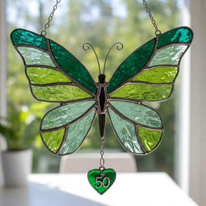 May include: Stained glass butterfly suncatcher in shades of green, with a heart-shaped charm that reads "50". The butterfly has a dark metal frame and is suspended by a chain. The wings feature various shades of green glass.