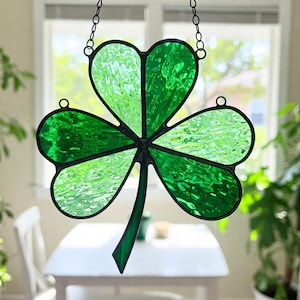 May include: A stained glass shamrock with a green and white color scheme. The shamrock is hanging from a chain and has a textured glass design.